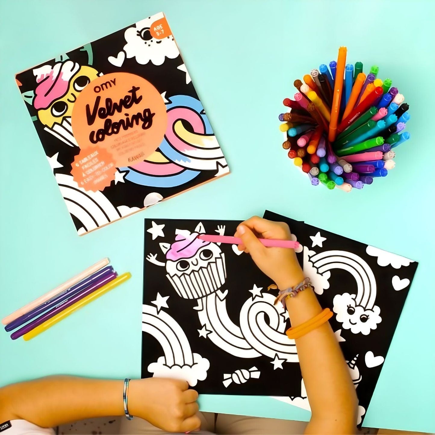 Criança a colorir o Livro colorir em veludo Lily da Omy com ilustrações kawaii, nuvens, arco-íris e cupcake sobre fundo colorido.
