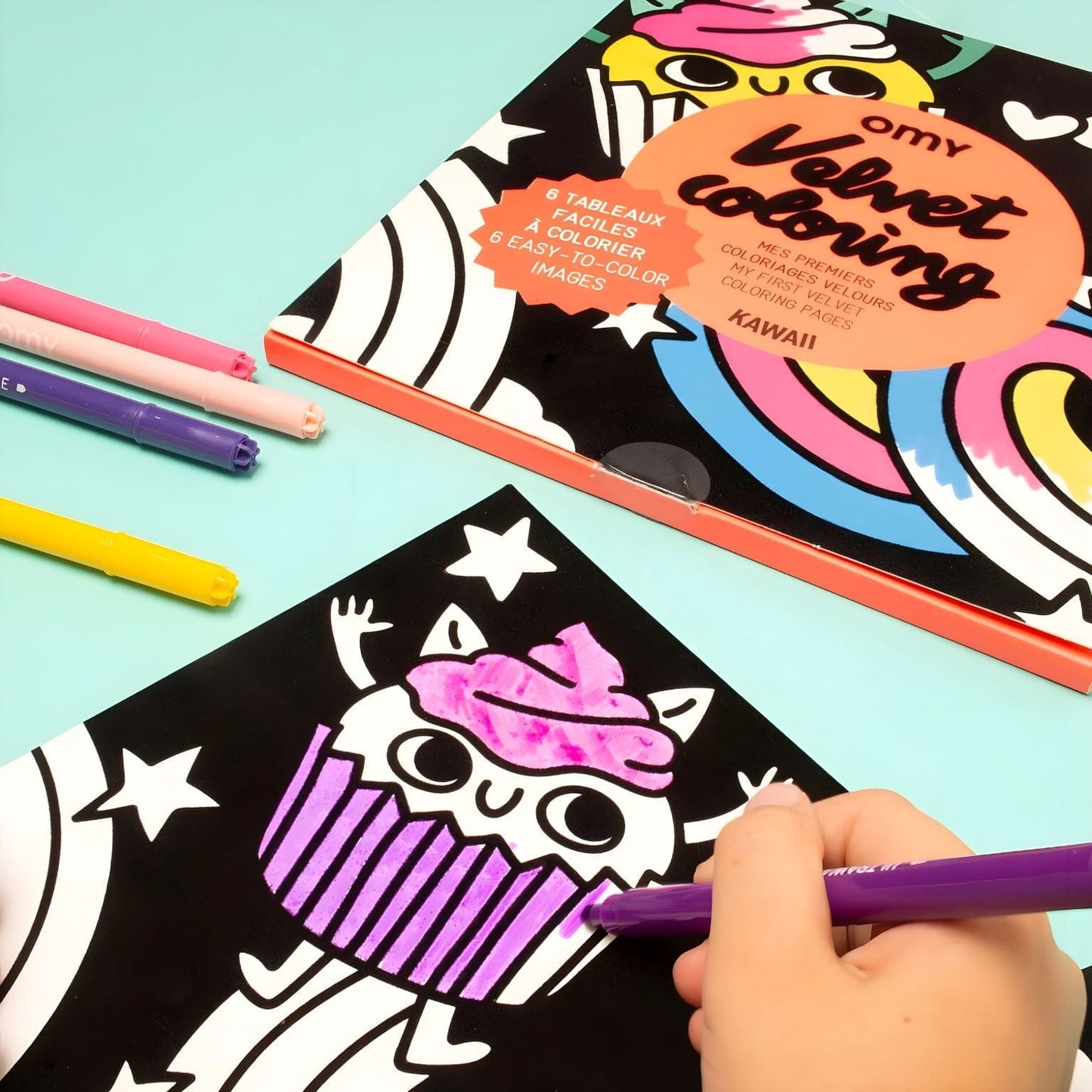 Criança a colorir o Livro colorir em veludo Lily da Omy com ilustrações kawaii, nuvens, arco-íris e cupcake sobre fundo colorido.